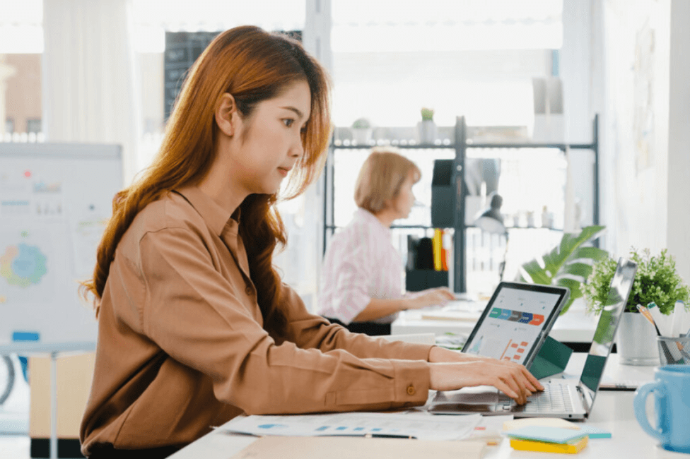 Mulher de cabelo ruivo e camisa bege sentada mexendo em um notebook