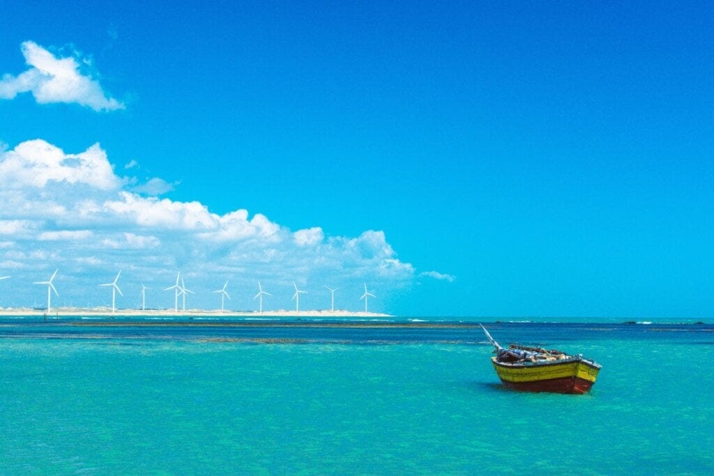 Praia de Icaraí de Amontada, com um mar azul-turquesa, um barco solitário flutuando e uma fileira de aerogeradores ao fundo