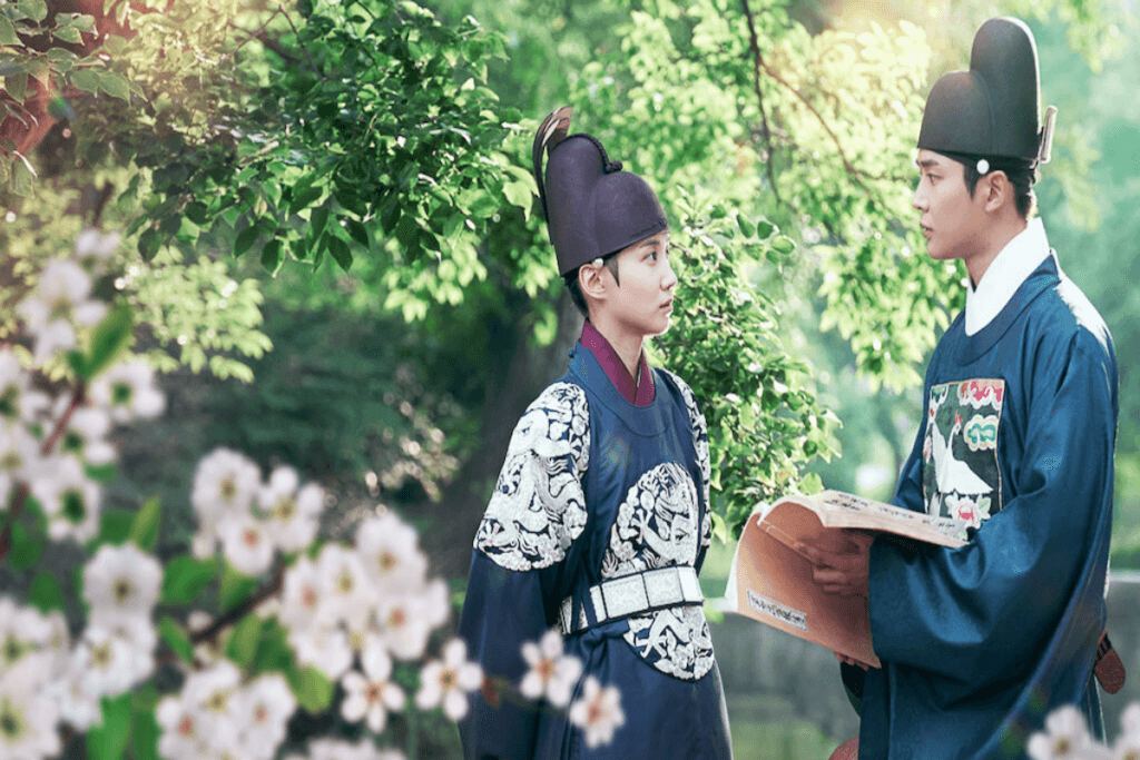 Dois homens conversando, um deles está com um livro na mão e ambos estão vestidos com roupas da era Joseon