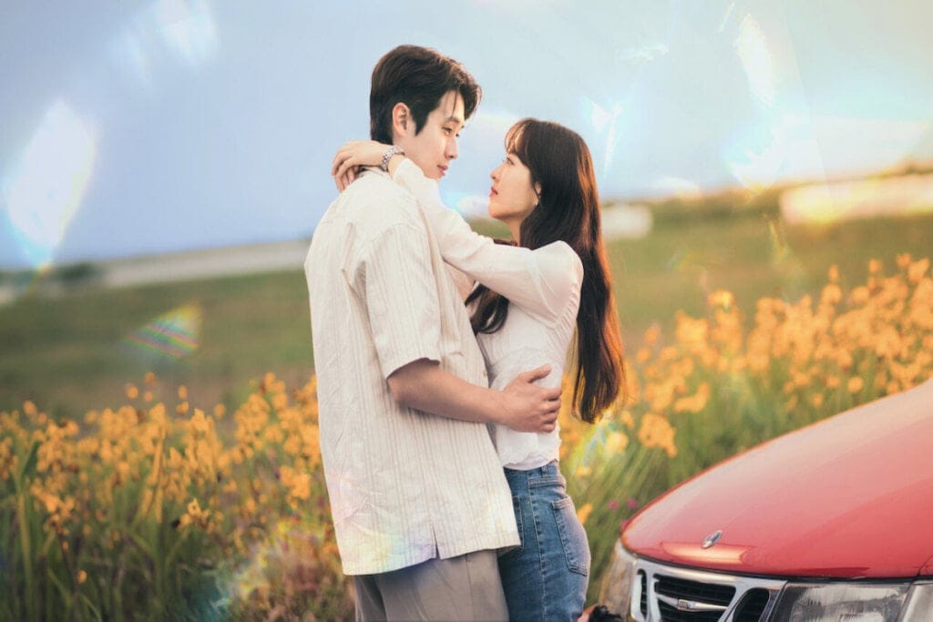Um casal se abraçando e atrás está um carro vermelho e um campo florido