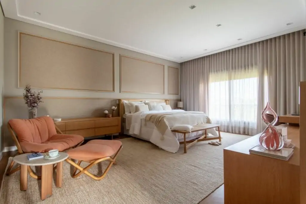 Dormitório com cama com forro branco e bege, tapete bege e janela lateral com cortinas bege