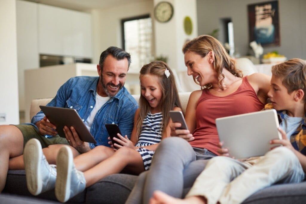 Pai, mãe e duas crianças com dispositivos eletrônicos na mão sentados no sofá  