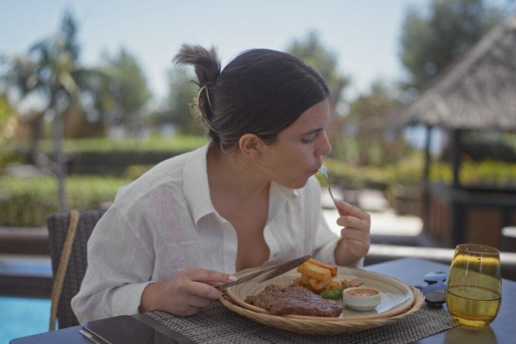 Mulher sentada de frente para uma vista com piscina comendo um bife
