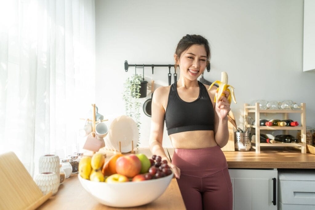 Jovem comendo banana na cozinha na bancada há uma cesta com frutas
