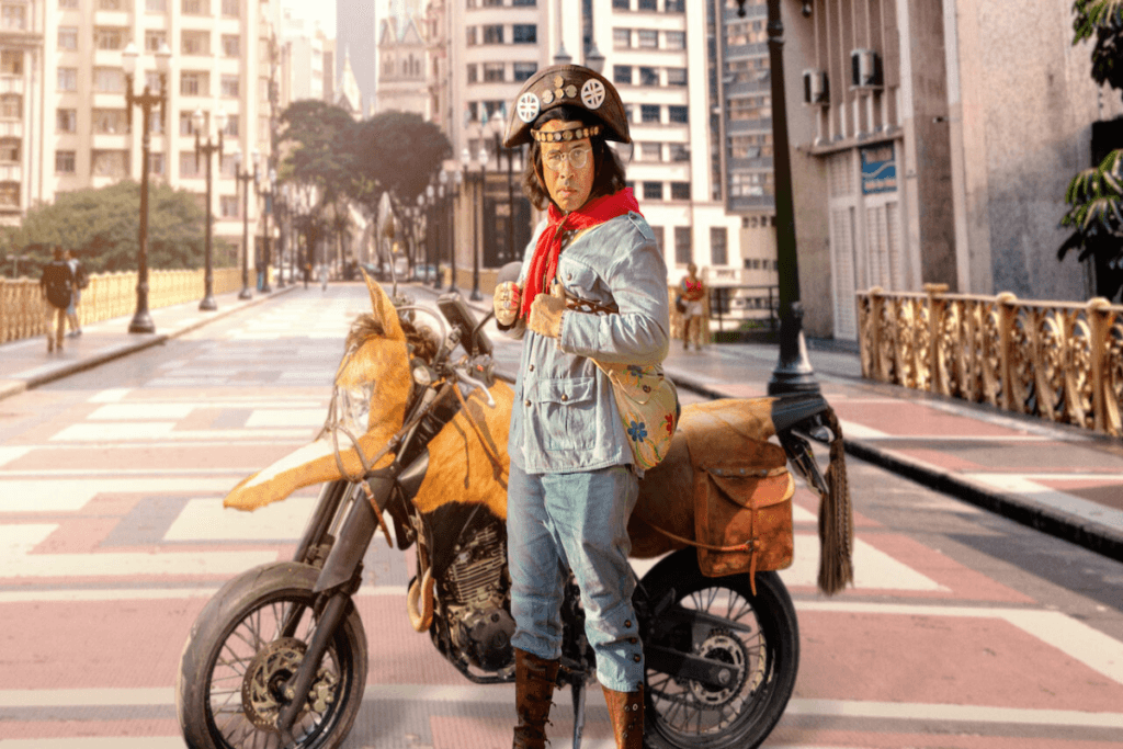Homem com roupa de cangaceiro, atrás dele esta uma moto e, no fundo, o centro de São Paulo