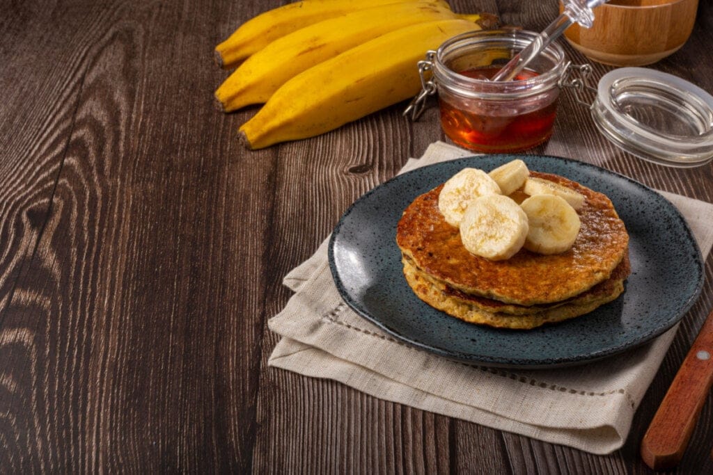 Prato preto com panqueca de banana, aveia e chia com banana e um pote de mel ao lado
