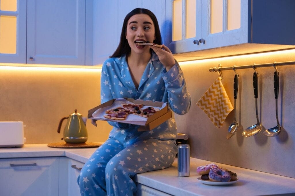 Mulher usando pijama e sentada na bancada de uma cozinha comendo pizza