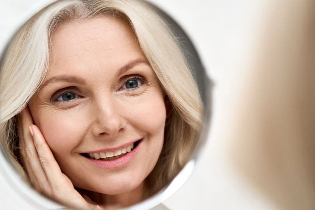 Reflexo de uma mulher sorrindo em um espelho e com uma das mãos no rosto 