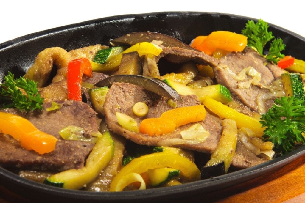 Recipiente preto com bifes, cebola, pimentões, abobrinha e berinjela 