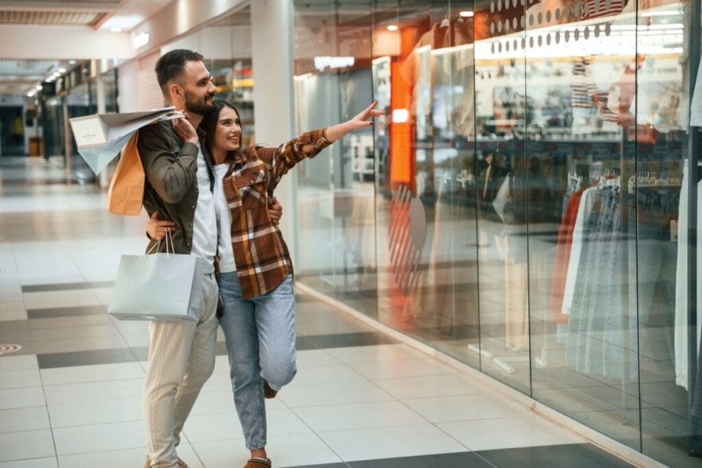 Casal abraçado e segurando sacolas de compras, olhando uma vitrine de loja