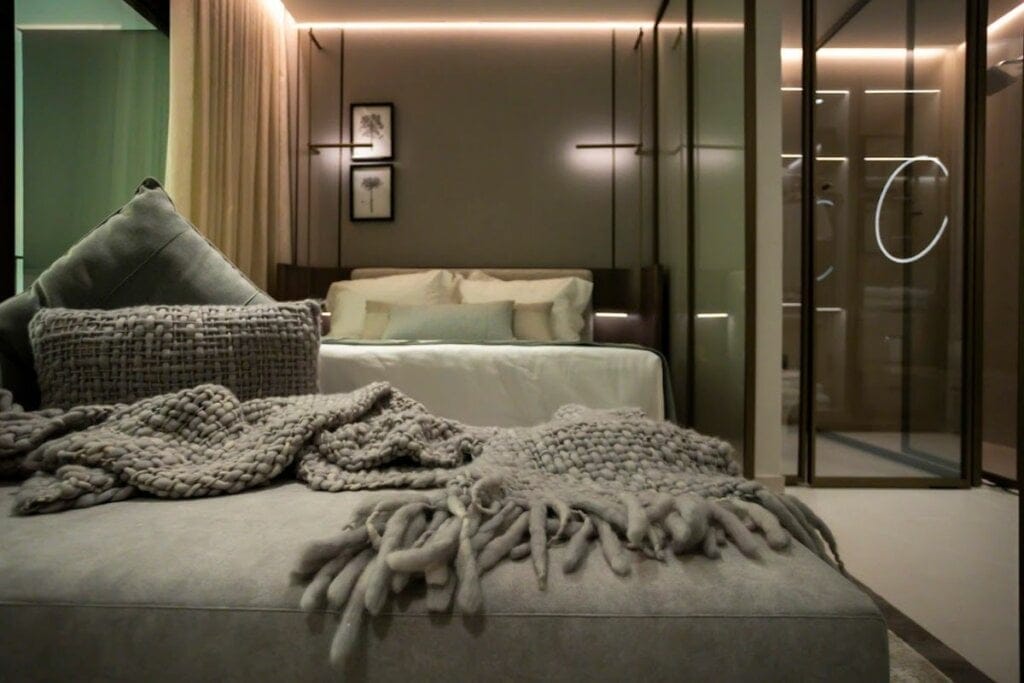 Imagem de um quarto com cama forrada ao fundo e uma poltrona cinza com manta da mesma cor na frente