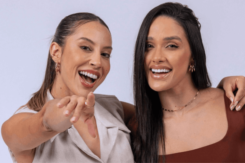 Duas mulheres, uma de cabelo preso com regata bege apontando para a câmera e a outra com o cabelo solto e com top marrom, elas estão abraçadas e sorrindo. 