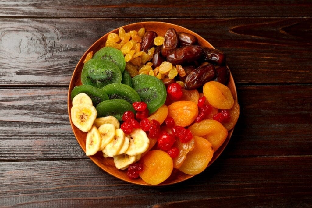 Prato com uma variedade de frutas secas, incluindo damascos, tâmaras, passas, kiwi desidratado, banana chips e cerejas, dispostos sobre uma mesa de madeira