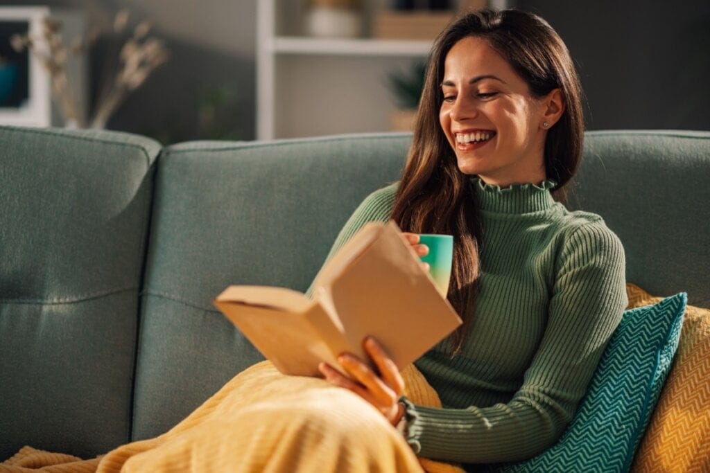 Mulher sorridente sentada em um sofá lendo um livro e segurando uma caneca