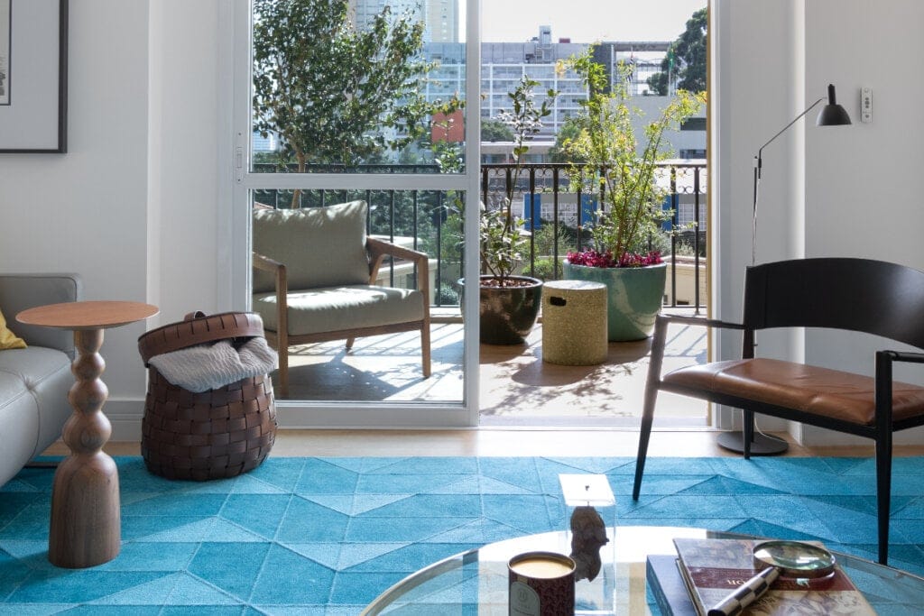 imagem de sala de estar com tapete grande azul, cadeira preta e porta de vidro ao fundo com varanda cheia de plantas