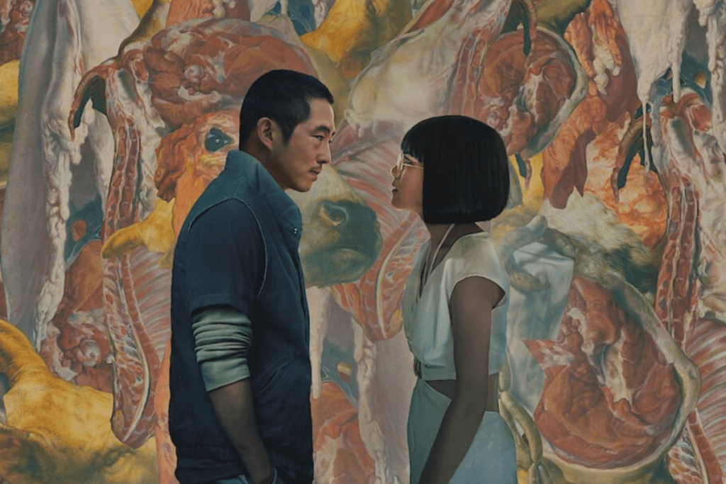 Cena da produção "Treta", com os protagonistas Steven Yeun e Ali Wong se encarando intensamente contra um fundo de imagens abstratas