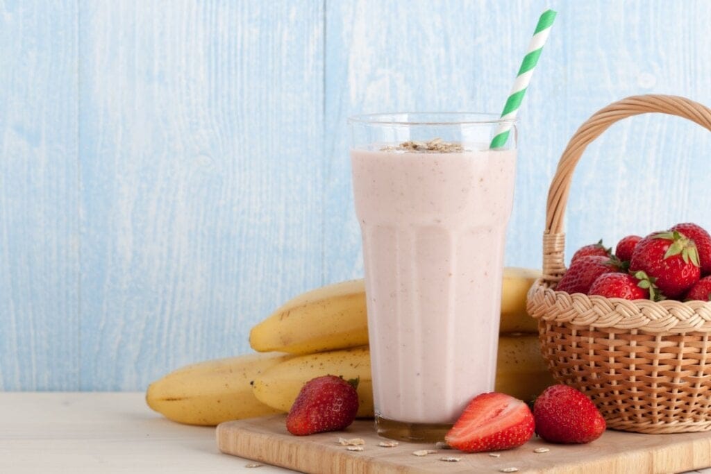 Copo de vidro com vitamina de morango com aveia e banana ao lado de uma cesta com morangos e bananas 